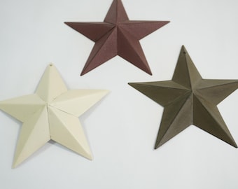 May include: Three metal star decorations in cream, dark brown, and olive green. Each star has a five-point design with a hole at the top for hanging. The stars are arranged on a white background.