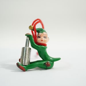 May include: A vintage Christmas ornament featuring a seated elf figure. The elf is dressed in a green outfit with a red collar and hat. It holds a silver object and has a red loop with a gold bell. The elf's face is painted with delicate features.