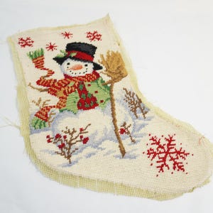 May include: A Christmas stocking with a cross-stitch design featuring a snowman wearing a black hat, red scarf, and green coat. The snowman is holding a broom in a snowy landscape with snowflakes.