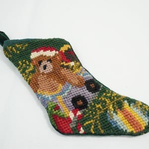 May include: A green Christmas stocking with a stitched design featuring a brown bear wearing a Santa hat. The stocking has a green loop for hanging and is decorated with colorful holiday imagery.