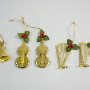 May include: A collection of gold-colored Christmas ornaments in the shape of musical instruments. The ornaments include a trumpet, two violins, and two harps. Each ornament is decorated with green leaves and red berries.
