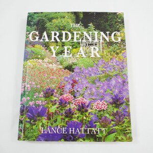 以下が含まれることがあります： ランス・ハッタット著「The Gardening Year」というタイトルの園芸本。表紙には、紫、ピンク、赤の花が咲き乱れる鮮やかな庭の風景が描かれています。タイトルは白で、装飾的な門のデザインが施されています。