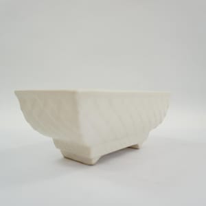 May include: An off-white ceramic planter, rectangular in shape, with a textured, diagonal pattern on the sides. It has a low, footed base. Suitable for small plants or succulents.