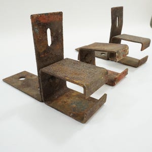 May include: Four weathered, rusty metal brackets with a rectangular base and a vertical support with an oval hole. The brackets have a U-shaped design, with a horizontal section and a short, angled lip. The metal has a mottled brown and orange appearance.