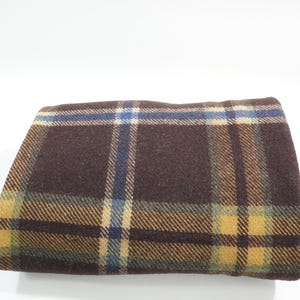 May include: Folded brown plaid fabric with blue and yellow accents. The plaid pattern features a repeating grid design. The fabric appears to be a wool or wool blend, and is folded over on itself.
