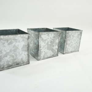 May include: Three galvanized metal square planters. The planters are a light gray color with visible weld lines and a textured surface. The open tops of the planters are visible. The planters are suitable for indoor or outdoor use.