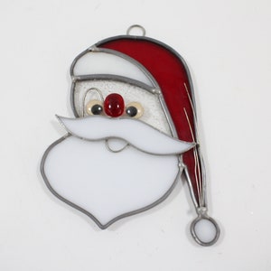May include: A stained glass ornament depicting Santa Claus's face. The ornament features a white beard, red hat, and black eyes with a red nose.