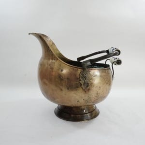 May include: Antique brass coal scuttle with a curved spout and a round base. The scuttle has a decorative metal emblem and a handle with white and blue ceramic accents. The coal scuttle is a vintage item.