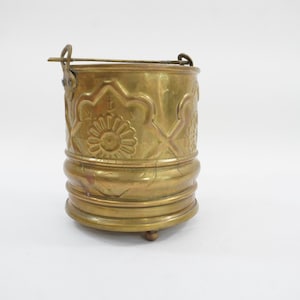 May include: A cylindrical brass bucket featuring a decorative embossed floral pattern around the upper section, a horizontal ribbed band near the base, and a metal swing handle attached to two side brackets with small feet at the bottom.