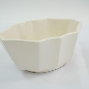 May include: A cream-coloured, geometric ceramic bowl with a faceted design. The bowl has a wide, open top and a slightly flared base. The interior is smooth, and the exterior has a matte finish. The bowl is suitable for serving or display.