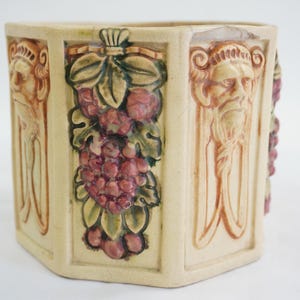May include: A beige ceramic planter with a hexagonal shape. The planter features relief carvings of a bearded face, grapes, and leaves in shades of red, green, and brown. The planter is a decorative item.