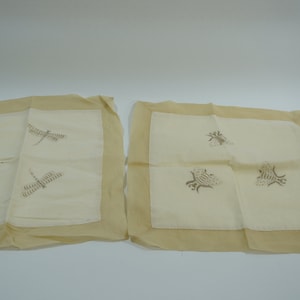 May include: Two square throw pillow covers with embroidered designs. One cover features three dragonfly designs in a light brown colour on a white background. The other cover features three bee designs in a light brown colour on a white background. Both covers have a light brown border.