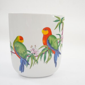 May include: A white ceramic mug featuring a colorful illustration of two parrots perched on a branch with green leaves and small pink flowers. The parrot on the left is primarily green and yellow, while the parrot on the right is red and green.