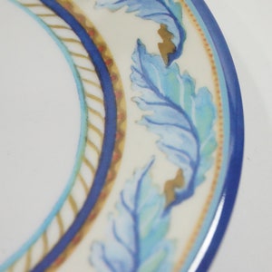 Może przedstawiać: Biały ceramiczny talerz z niebiesko-złotym wzorem liści na obrzeżu. Talerz ma niebieską obwódkę i kremowy wewnętrzny pas z wzorem przypominającym linę. Liście są w odcieniach niebieskiego i złotego.