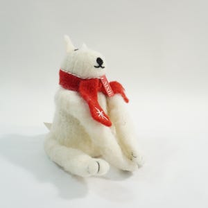 May include: A white felted dog figurine with a red scarf. The scarf has a white star and the word "NIXIE" printed on it. The dog is sitting with its legs extended and its ears perked up.