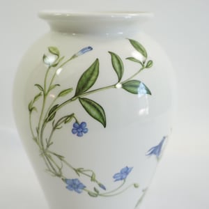 May include: A white ceramic vase with a rounded body and a narrow neck. The vase is decorated with a delicate floral pattern of green leaves and small blue flowers. The design is painted in a realistic style.