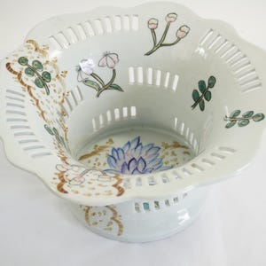 May include: A white ceramic basket with a scalloped rim and a pedestal base. The basket features a pierced design and is decorated with hand-painted floral and leaf motifs in shades of blue, pink, green, and brown.
