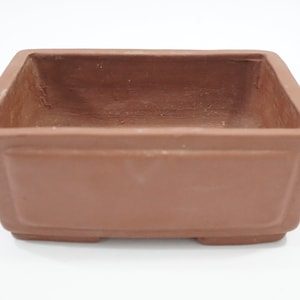 以下が含まれることがあります： テラコッタ色の正方形の盆栽鉢。鉢は長方形で、わずかに先細りのデザインと隆起したベースがあります。内部が見え、鉢の深さを示しています。