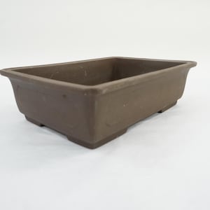 Puede incluir: Una maceta rectangular marrón para bonsái con una superficie ligeramente texturizada. La maceta tiene un diseño simple y clásico con un borde elevado y pequeños pies. Ideal para bonsáis y otras plantas pequeñas.