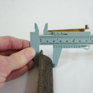 May include: A close-up of a metal decorative piece with intricate details. The piece is being measured with a caliper, showing a measurement of 6.8 cm.