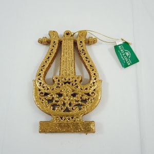 May include: Gold glitter lyre ornament with a Kurt S. Adler tag. The lyre is a musical instrument with a decorative design.