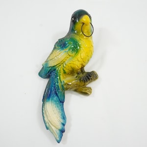 May include: A colorful parrot figurine, perched on a branch. The bird features a yellow body, blue and green wings, and a blue tail. A black metal ring is attached to the head. The figurine is likely made of ceramic or a similar material.