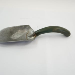 May include: A small, vintage metal scoop with a curved, dark green handle. The scoop has a worn, silver finish, suggesting age and use. The handle is smooth and ergonomically designed for a comfortable grip. This tool is likely used for gardening or other small tasks.