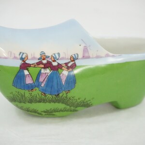 May include: A ceramic Dutch clog-shaped bowl, painted green with a scene of figures in traditional attire dancing in a circle. The background features a blue sky, water, and windmills.