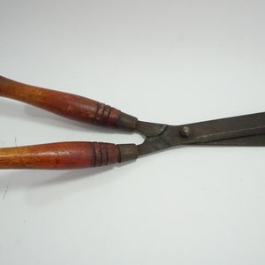 Op de afbeelding: Een heggenschaar met houten handvatten en donkere metalen bladen. De handvatten zijn warmbruin van kleur en de bladen zijn van donkerder, verouderd metaal. De schaar is ontworpen voor het trimmen en vormen van heggen en struiken.