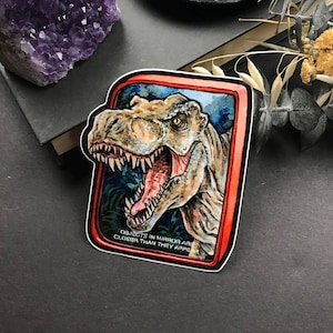 May include: A sticker featuring a detailed illustration of a Tyrannosaurus Rex with an open mouth, showcasing sharp teeth and a red tongue. The dinosaur is set against a blue and green background within a red-bordered frame. The text "OBJECTS IN MIRROR ARE CLOSER THAN THEY APPEAR" is at the bottom.