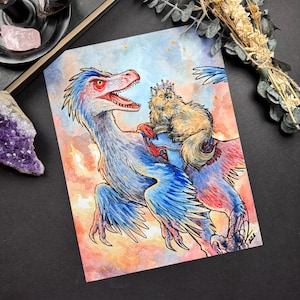Puede incluir: Impresión artística en acuarela que presenta un dinosaurio azul y rojo con un gato con corona montado en su espalda. El fondo tiene un cielo colorido con tonos naranja y azul. El gato tiene pelo largo.