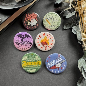 Könnte beinhalten: Eine Sammlung runder Ansteckbuttons mit verschiedenen Designs und Texten. Die Buttons zeigen Themen wie Horror, Fantasy und Romantik, mit Illustrationen eines Schädels, eines Drachen und einer Burg. Der Text enthält "Horror Reader", "Fan Fic reader" und "Thriller Reader".