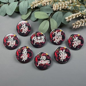 Peut inclure: Huit boutons ronds avec différentes options de pronoms imprimées dessus. Les boutons sont noirs avec un motif floral rouge et un texte blanc. Les options incluent : "She They", "He She They", "Ask me my pronouns", "She Her", "They Them", "He Him", et "Any pronouns".