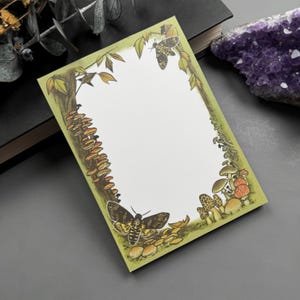May include: A rectangular notepad with a nature-themed illustration. The border features a green frame with mushrooms, trees, and moths. The illustration includes a large moth at the bottom and a smaller one at the top. The notepad has a blank white space for writing.