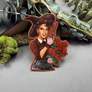 May include: A woman with brown hair wearing a witch's hat and a brown dress holds a bouquet of red roses and a pair of scissors.