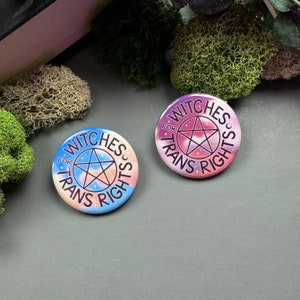 Peut inclure: Deux boutons ronds avec un fond dégradé rose et bleu. Les deux boutons présentent un pentagramme avec un contour blanc et un remplissage noir. Le texte sur les boutons indique "Witches for Trans Rights".