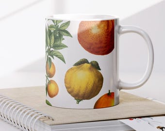 May include: White ceramic mug featuring a botanical illustration of oranges, lemons, and other citrus fruits. The mug is set on a spiral-bound notebook. The design includes green leaves and various orange and yellow fruits.