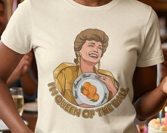 May include: A woman wearing a cream-colored t-shirt with a cartoon image of a woman holding a plate of oranges. The text on the shirt reads "I'M QUEEN OF THE BALL". The background includes balloons, a birthday cake, and people.