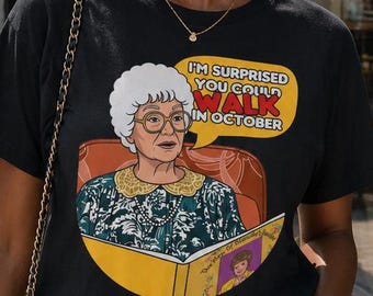 May include: A black t-shirt featuring a cartoon illustration of an older woman reading a book, with the text "I'M SURPRISED YOU COULD WALK IN OCTOBER" in a speech bubble. The person is wearing sunglasses and blue jeans.