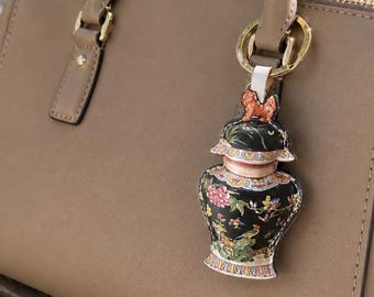 May include: A structured beige handbag with dual top handles and a gold-toned metal ring attachment. Hanging from the ring is a decorative charm shaped like a black ginger jar, featuring a colorful floral pattern and a small orange animal figure on top.