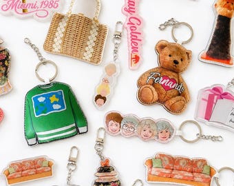 May include: A collection of acrylic keychains featuring various designs including a palm tree with the text Picture it! Miami, 1985, a wicker bag, a teddy bear with the name Fernand, a green sweater, a floral vase, a sofa, and portraits of four women.