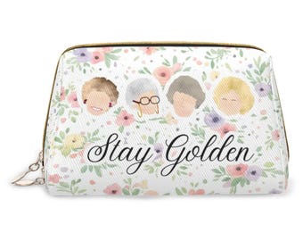 May include: A white cosmetic pouch featuring a floral pattern in pastel pink, purple, and green. The design includes four stylized character heads and the black cursive text Stay Golden. The bag has a gold-toned zipper closure along the top edge.