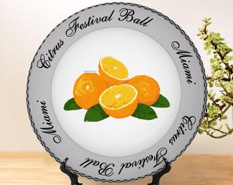 May include: A white ceramic plate featuring a central illustration of bright orange citrus fruits with green leaves. The gray border displays the repeating black text Citrus Festival Ball Miami in a cursive font with a checkered edge pattern.