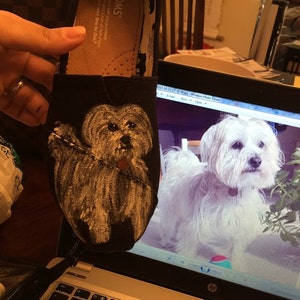 Könnte beinhalten: Ein schwarzer Canvas-Schuh mit einem handgemalten Bild eines weißen Hundes. Der Schuh ist ein Schuh der Marke TOMS. Der Schuh wird in der Hand einer Person gehalten. Der Schuh befindet sich neben einem Laptop-Bildschirm, auf dem ein Foto eines weißen Hundes zu sehen ist.