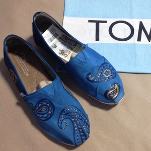 Könnte beinhalten: Ein Paar blaue Canvas-Schuhe mit goldenen Paisley-Mustern. Die Schuhe befinden sich auf einer braunen Oberfläche.