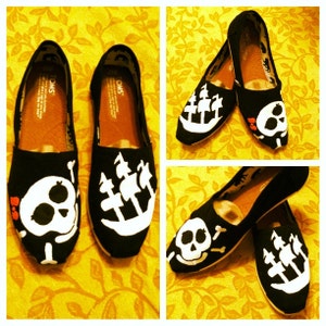 Könnte beinhalten: Ein Paar schwarze Canvas-Schuhe mit weißen, bemalten Designs. Ein Schuh hat ein Totenkopf- und Knochen-Design, der andere ein Schiff mit drei Masten. Die Schuhe haben das Wort "TOMS" an der Seite.