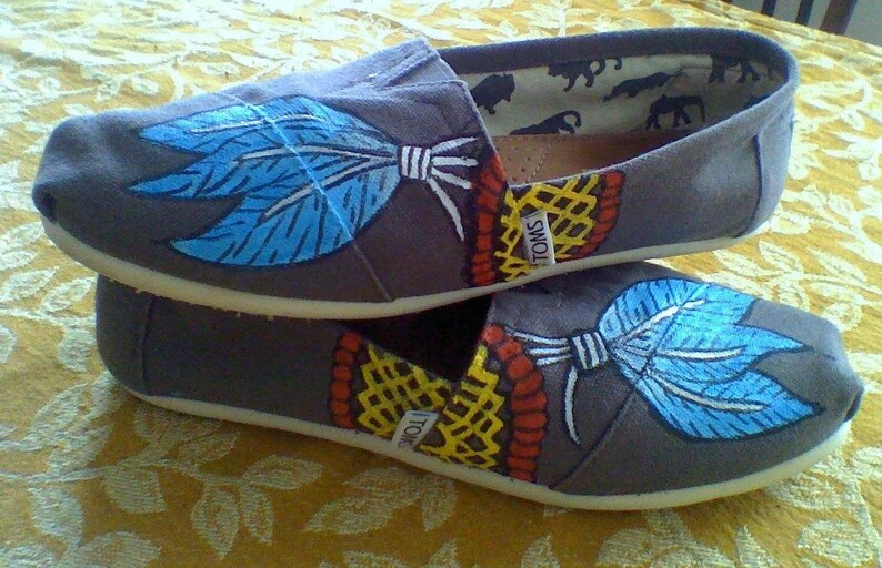 K&ouml;nnte beinhalten: Ein Paar graue Canvas-Schuhe mit blauen und wei&szlig;en gemalten Federn und einem geometrischen Muster in Gelb, Rot und Schwarz. Die Schuhe haben den Schriftzug "TOMS" aufgedruckt.