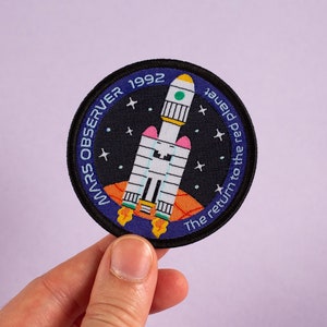 Könnte beinhalten: Ein blauer und schwarzer gestickter Aufnäher mit einer Rakete und Sternen. Der Text auf dem Aufnäher lautet "Mars Observer 1992 The return to the red planet".