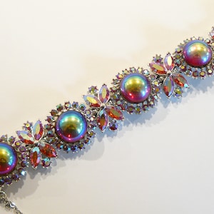 Könnte beinhalten: Ein silbernes Armband mit schillernden roten, grünen und blauen Steinen. Die Steine sind in einem floralen Design eingefasst und von kleineren klaren Steinen umgeben.