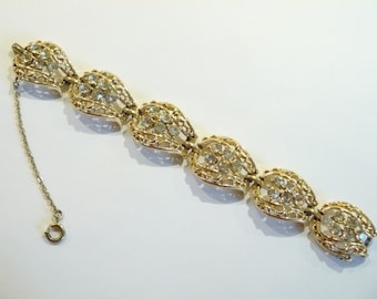 Puede incluir: Un brazalete de tono dorado con cierre de cadena. El brazalete está hecho de una serie de piezas enlazadas, ornamentadas, en forma de lágrima, cada una engastada con diamantes de imitación transparentes.